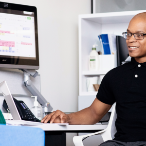 A business man works on a laptop with a monitor showing a spreadsheet in the background