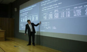 A professor stands in front of a large screen of numbers