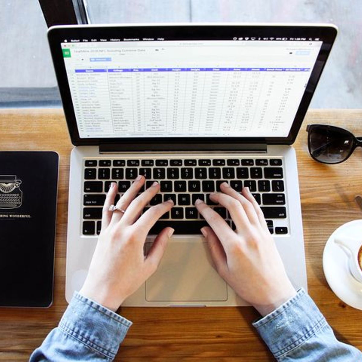 Hands typing on a laptop. A spreadsheet is visible on the screen.