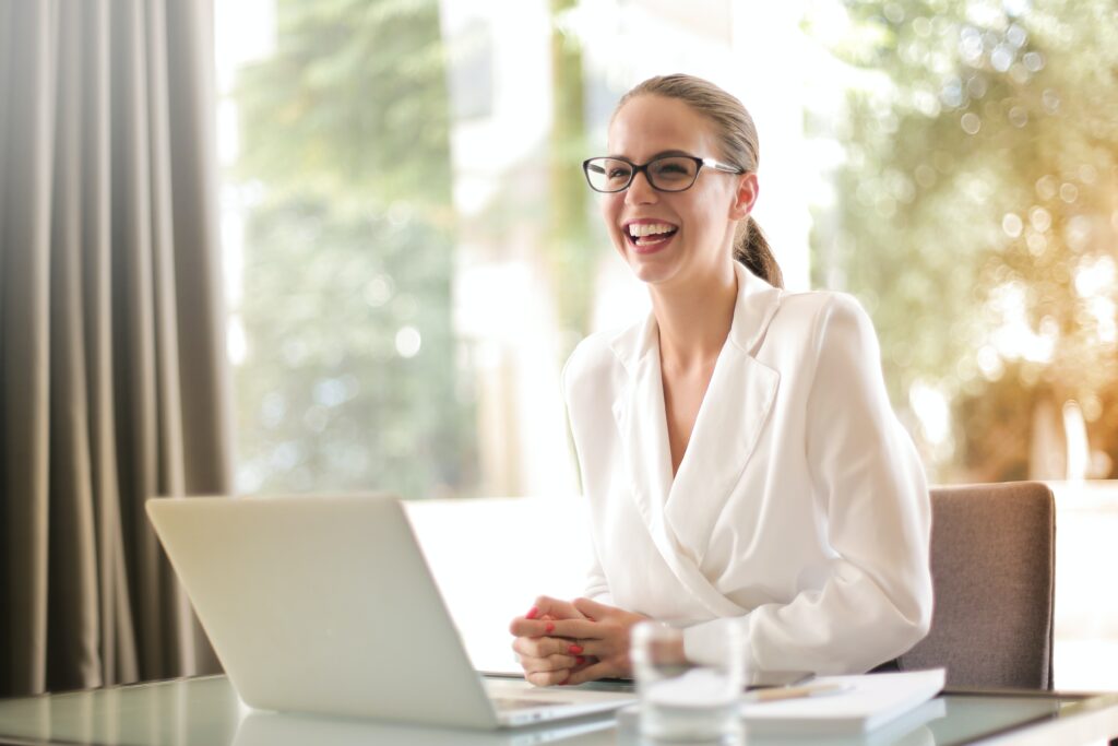 A woman in business attire at a desk with an open laptop. The woman is looking offscreen and laughing.
