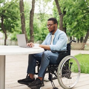 A man in a wheelchair sits outside at a table. He is working on a laptop. A cup of coffee is next to the laptop.